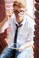 Using opportunities of digital age. Top view of handsome young man in shirt and tie working on digital tablet and looking at camera while sitting at the window sill photo