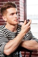 Smoking pipe. Handsome young man in striped shirt smoking pipe and looking away while sitting on the chair photo