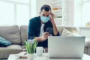 Young man in protective face mask using smart phone while working from home photo