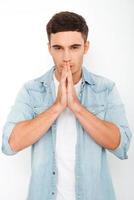 Begging for... Thoughtful young man holding hands clasped near face and looking at camera while standing against white background photo