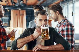 Charming young man in casual clothing drinking beer while spending time with friends in the pub photo