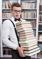 llevando una pila de libros. joven conmocionado con camisa y corbata de moño que lleva una pila de libros pesados y mira la cámara foto