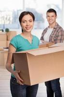 Unpacking boxes. Attractive young woman holding an opened cardboard box and smiling at camera while cheerful man standing on background photo