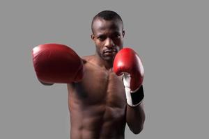 boxeador confiado. confiado joven boxeador africano en guantes de boxeo estirando su mano y mirando a la cámara mientras está de pie aislado en gris foto