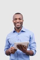 Working on digital tablet. Front view of handsome African man working on digital tablet and smiling at camera while standing isolated on grey photo