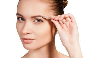 Tweezing her eyebrows. Beautiful young shirtless woman tweezing her eyebrows and looking at camera while standing isolated on white background photo