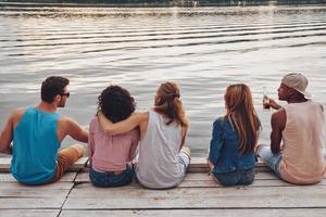 Doing whatever they want. Rear view of young people in casual wear talking while sitting on the pier photo