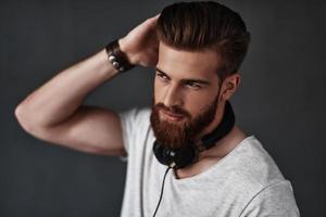 Calm and confident.  Handsome young man wearing headphones around his neck and running his hand through his hair while standing against grey background photo