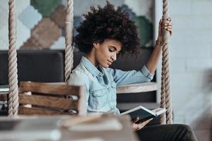 disfrutando de un buen libro. bella joven africana leyendo un libro mientras se sienta en el columpio foto