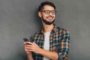 siempre en contacto un joven alegre con anteojos sosteniendo su smartphone y mirando hacia otro lado con una sonrisa mientras se enfrenta a un fondo gris foto