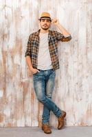 Serious and confident. Full length of handsome young man adjusting his hat and looking at camera while standing against the wooden wall photo