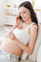 Calling doctor for consultation. Beautiful pregnant woman sitting on sofa and talking on the mobile phone while looking at the paper photo