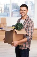 Just moved in a new house. Handsome young man holding an cardboard box with home stuff in it and smiling while more carton boxes laying on background photo