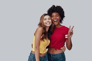 dos mujeres jóvenes atractivas mirando a la cámara y sonriendo mientras están de pie contra el fondo gris foto