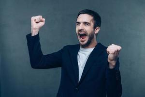 Happy winner. Happy young man gesturing and keeping his mouth open while standing against grey background photo