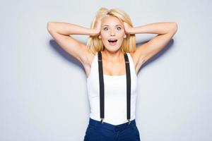 Shocked and happy. Surprised young woman holding hands in hair and staring at camera while standing against grey background photo