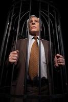 Men in cell. Low angle view of rustrated senior man in formalwear standing in cell and looking away while isolated on black background photo