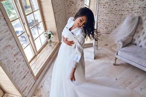 Enjoying her special day. Top view of attractive young woman in silk bathrobe trying on her wedding dress and smiling while standing near window