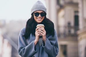 The weather is great today Beautiful young woman in sunglasses holding coffee cup and looking at camera with smile while standing outdoors photo