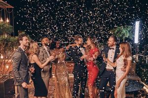 Group of happy people in formalwear having fun together with confetti flying all around photo