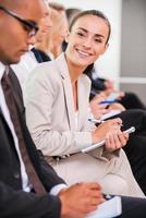 Businesswoman at the conference. Side view of business people sitting in a row and writing something in their note pads while confident young woman looking at camera and smiling photo