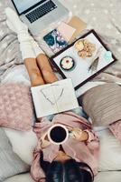Top view of beautiful young woman in pajamas enjoying morning coffee while resting in bed at home photo