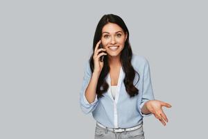 buena plática. mujer joven sorprendida hablando por teléfono mientras está de pie contra el fondo gris foto