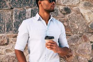 hombre disfrutando de café recién hecho. primer plano de un apuesto joven africano sosteniendo una taza de café mientras está de pie contra la pared empedrada al aire libre foto