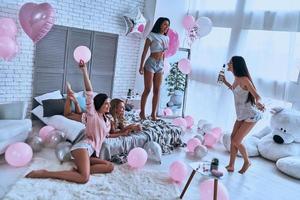 ¿Quién quiere un trago? Cuatro atractivas mujeres jóvenes en pijama uniéndose mientras hacen una fiesta de pijamas en el dormitorio con globos por todas partes. foto