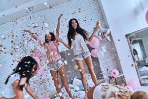 So much fun Four attractive young women in pajamas smiling and gesturing while jumping in the bedroom with confetti flying everywhere photo