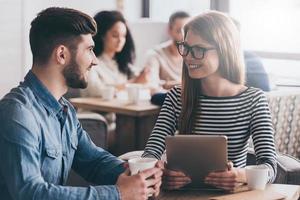 Sharing good news. Two people looking at each other with smile while sitting at the office table with people at the background photo