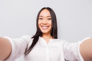 Happy moments must be saved. Cheerful young Asian woman holding camera and making selfie while standing against grey background photo