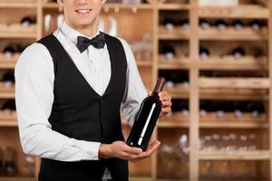 Presenting the best wine. Cropped image of cheerful young sommelier standing in front of wine shelf and holding a wine bottle photo