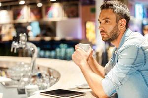 Coffee for inspiration. Side view of young man holding cup of coffee while sitting at the bar counter photo