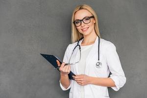 Hello I am your doctor. Beautiful young female doctor holding clipboard and looking at camera with smile while standing against grey background photo