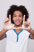 Finger frame. Cheerful African teenager looking through a finger frame and smiling while standing isolated on grey background photo