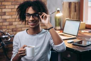 descanso. alegre joven africano sosteniendo una taza de café y ajustando sus lentes con una sonrisa mientras se sienta al lado de su lugar de trabajo foto