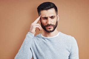 Frustrated young man in casual wear touching his head with hand while standing against brown background photo