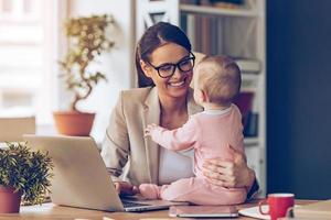 Working together is so fun  Cheerful young beautiful businesswoman looking at her baby girl with smile while sitting at her working place photo