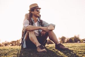 What a beautiful day Handsome young man in fedora carrying backpack and smiling while sitting on green grass outdoors photo