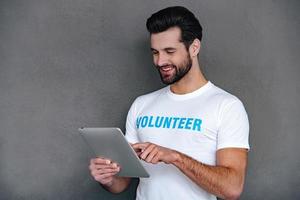 Donation by one click. Confident young man in volunteer t-shirt using his digital tablet with smile while standing against grey background photo