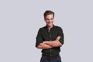 Too good to be real. Good looking young man keeping arms crossed while standing against grey background photo