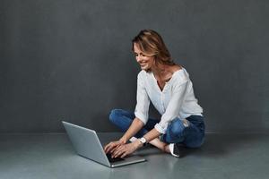 Shopping online. Attractive young woman in casual wear using computer and smiling while sitting on the floor against grey background photo