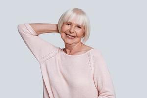 simplemente feliz. hermosa anciana mirando a la cámara y sonriendo mientras está de pie contra el fondo gris foto