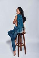 Feeling free and confident. Full length of attractive young woman carrying on shoulders denim jacket while sitting on the chair against grey background photo