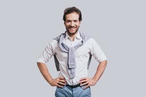 Young and successful. Beautiful young man in smart casual clothes keeping hands on hip and looking at camera while standing against grey background photo