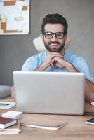 Great solution. Cheerful young handsome man wearing glasses looking at camera with smile while sitting at his working place photo