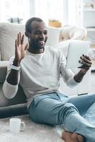 Pleasant talk with friend. Handsome young African man talking with his friend using digital tablet while sitting on the carpet at home photo