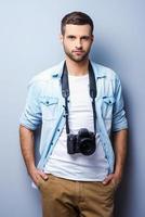 Confident and experienced photographer. Handsome young man with digital camera looking at you while standing against grey background photo