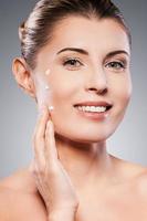 Keeping her skin young and smooth. Portrait of shirtless mature woman spreading cream on face and smiling while standing against grey background photo